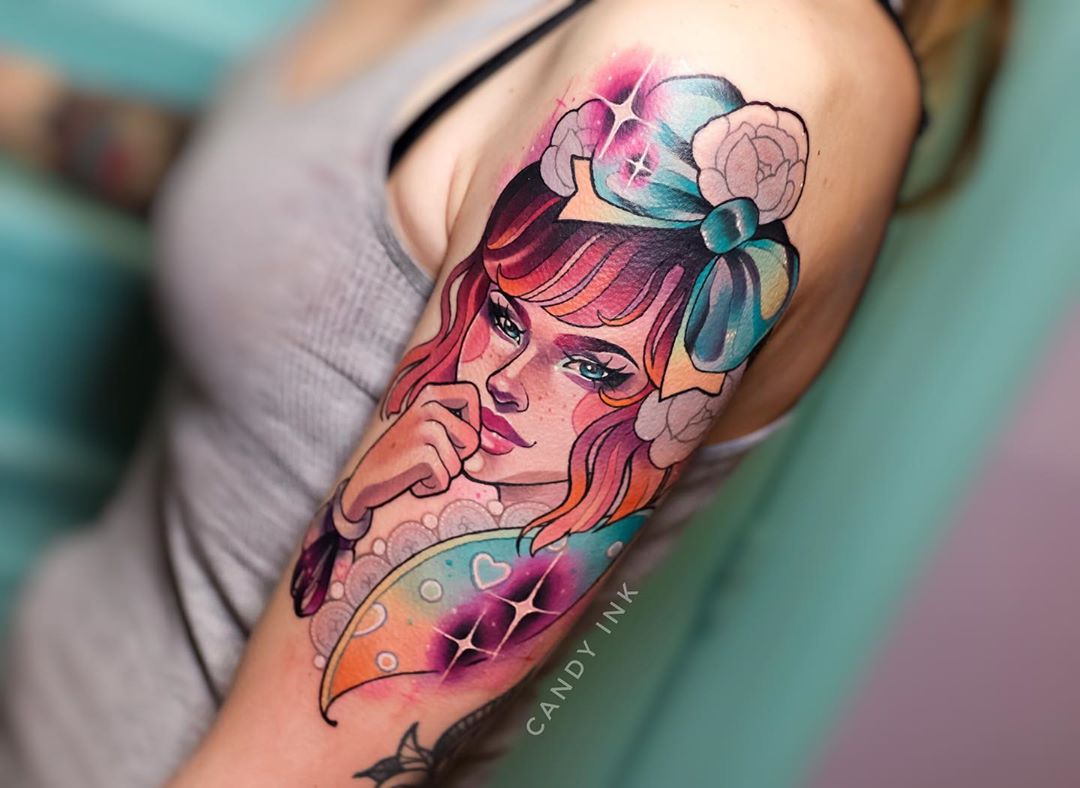 a closeup photo showcasing a colorful neotraditional tattoo of a woman on the upper arm. the woman has red hair, blue eyes, and a blue bow in her hair. there are also flowers and sparkles incorporated into the tattoo. the tattoo artist's signature, "candy ink", is visible on the inner bicep.