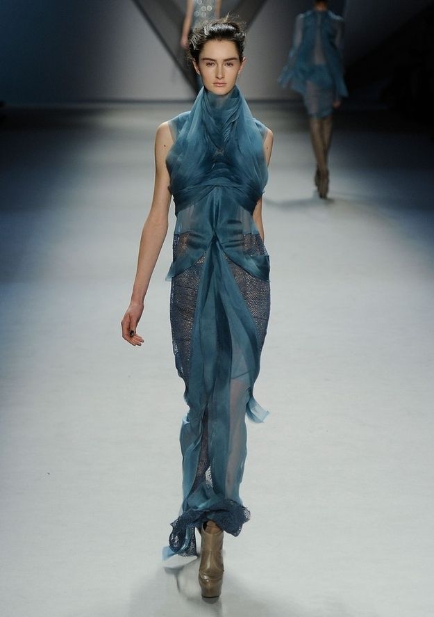 a female model walks the runway wearing a long, flowy blue gown. her hair is swept back and she looks straight ahead with a confident expression. the runway is bare and white, allowing the dress to stand out. the overall impression is one of elegance and sophistication.