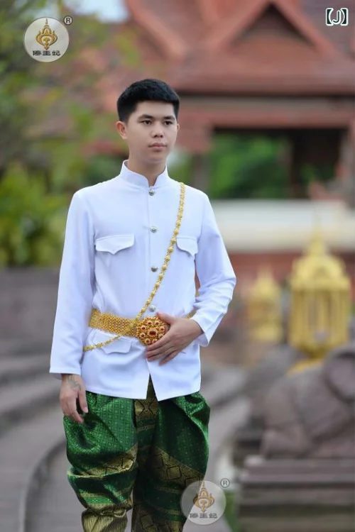 a man in traditional thai wedding attire is standing outside a temple. he is wearing a white longsleeved shirt with a high collar, green pants, and a gold belt. he is smiling and looks very happy. the temple in the background is decorated with gold accents, creating a beautiful setting for the photo.