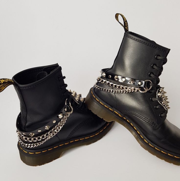 the image shows a pair of black dr. martens boots adorned with multiple silver chains and studded accents. the chains wrap around the ankles and incorporate small, pyramidshaped studs. the boots themselves are classic dr. martens style with yellow stitching on the soles and prominent studs along the sides.  the overall aesthetic is punk or grungeinspired, showcasing a rebellious and edgy fashion statement.