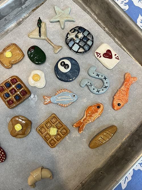 the image shows a variety of ceramic food and symbol decorations laid out on a baking tray lined with parchment paper. there are breakfast themed decorations like pancakes, waffles, eggs, a croissant, and fish. there are also symbol and object shaped decorations such as a starfish, a cocktail glass with an olive, an eight ball, an ace of hearts playing card, and a horseshoe.