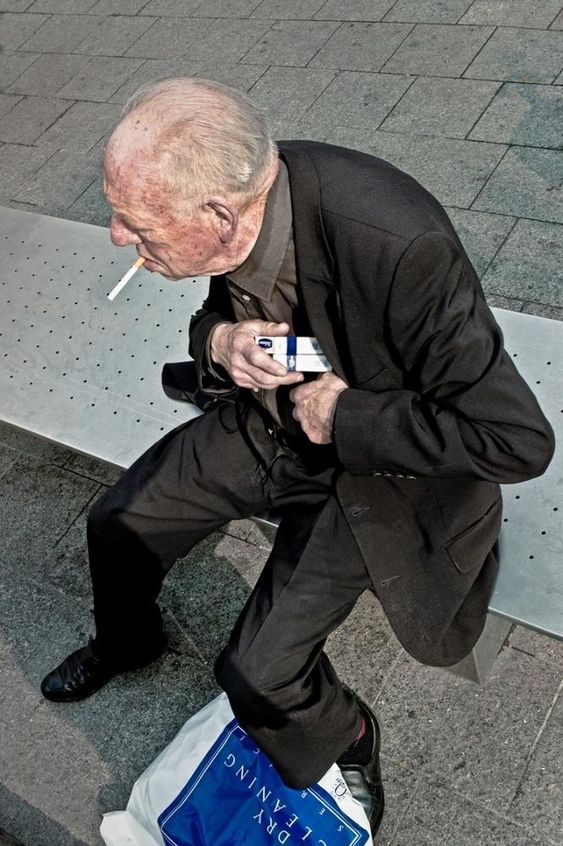 a highangle shot of an elderly man in a black suit sitting on a metal bench. he is smoking a cigarette and holding a cell phone in his other hand. the man is looking down, and his face is partially obscured. the setting appears to be an urban sidewalk or plaza. the overall tone of the image is candid and observational.