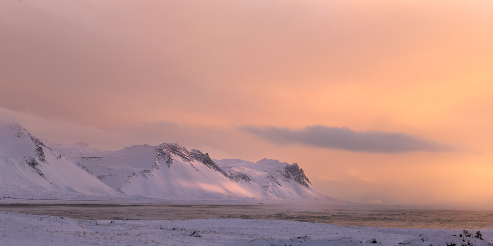 a snowy mountain range, the tops dusted with pink and orange hues from a beautiful sunset. the sky is a soft, pastel pink, with a few wispy clouds. the scene is peaceful and tranquil, with a vast, frozen wilderness stretching out in front of the viewer. the snow is pristine white, untouched by any footprints.