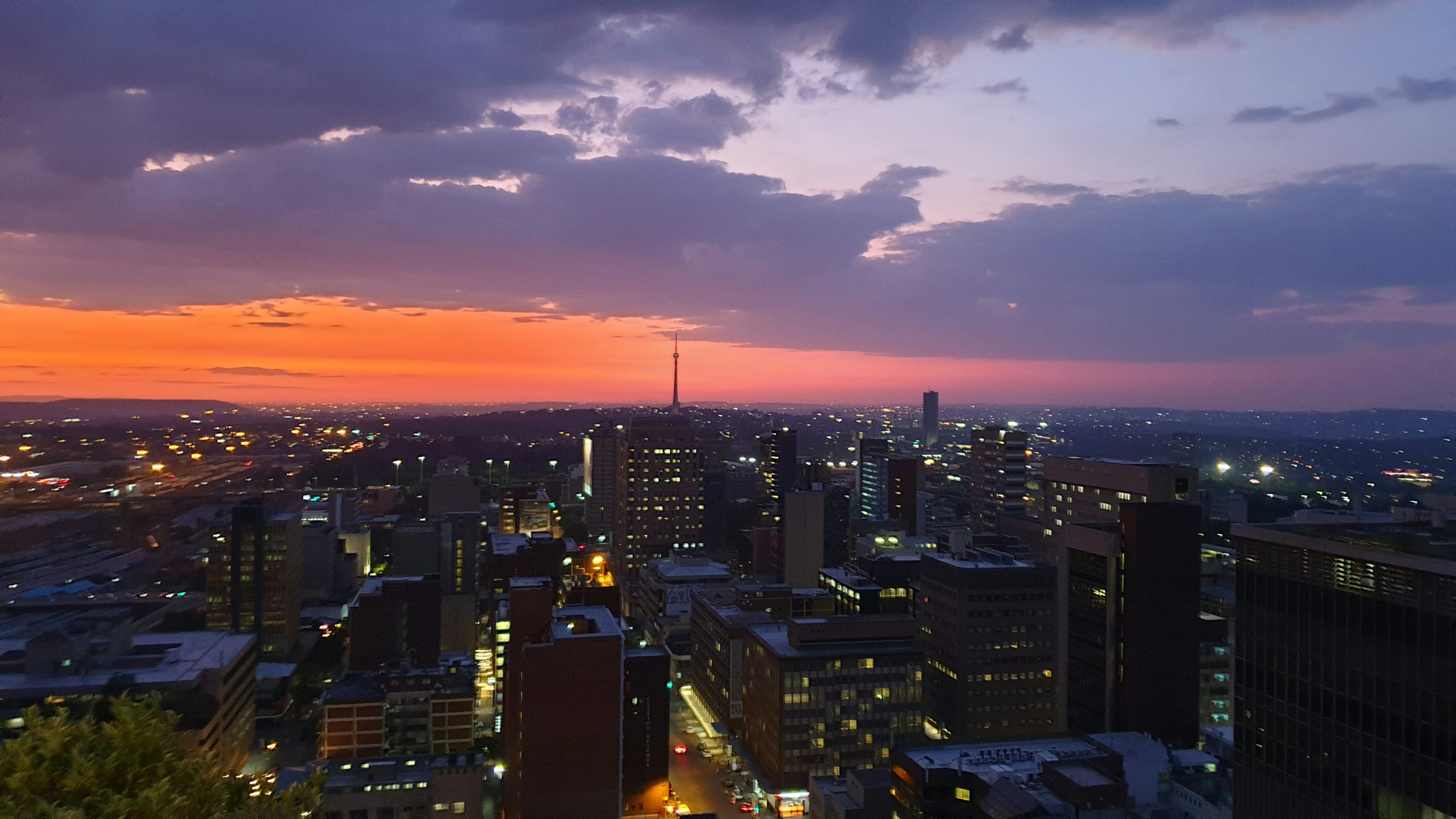 this is a wide angle shot of a cityscape at sunset or twilight. tall buildings and skyscrapers fill the foreground and middle ground, illuminated by city lights. in the background, the sky is a mix of vibrant orange, pink, and purple colors, with some scattered clouds. an antenna or tower rises above the skyline in the distance. the scene appears to be taken from a high vantage point, providing a panoramic view of the urban landscape. the location is likely johannesburg, south africa.