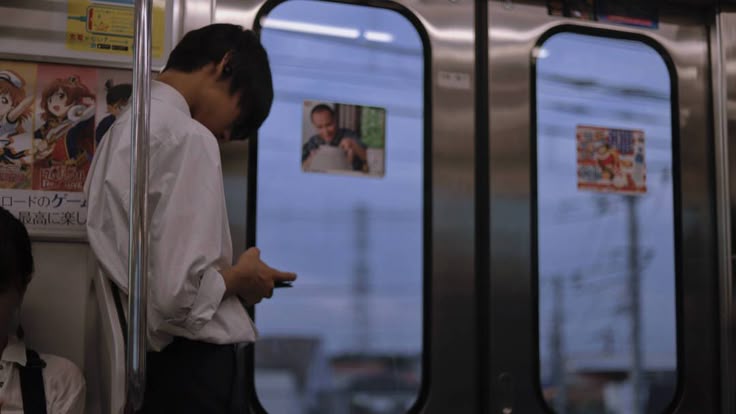 a young man stands on a commuter train in japan, looking down at his cell phone. he's wearing a white shirt and dark pants. the train car has advertisements featuring animestyle characters and blurry views of the passing cityscape outside the windows. the overall scene suggests a typical daily commute.