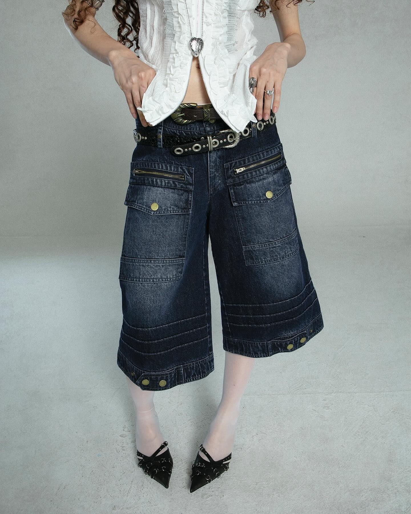 a fulllength shot of a woman modeling a stylish outfit. she's wearing a white, ruffled blouse tucked into a pair of darkwash denim culottes. the culottes feature multiple pockets, a detailed stitching design, and are secured with a dark belt featuring metallic accents. she's accessorized with rings and is wearing black heeled shoes. the background is a simple, light grey studio setting. the overall style evokes a vintage or retro feel, potentially reminiscent of 90s or y2k fashion trends.