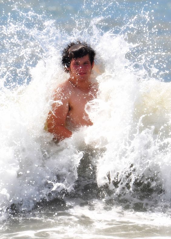a young man is standing in the ocean with waves crashing around him. he is smiling and appears to be enjoying the cool water on a warm day.