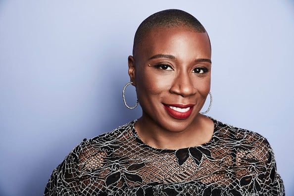 a portrait of a smiling black woman with a shaved head, hoop earrings, and red lipstick, wearing a black lace top against a blue background.