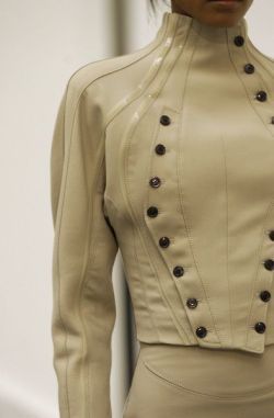 a closeup shot of a woman wearing a stylish, modern, creamcolored leather jacket. the jacket features a high neckline, long sleeves, and a series of buttons down the front. the model is wearing a matching skirt and the jacket is highlighted against a white background. the image captures the detail and craftsmanship of the jacket, showcasing its sleek and sophisticated design.