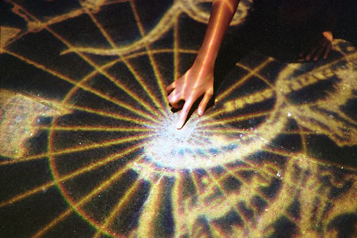 a hand with a finger extended towards the center of a starburstlike projection on a surface, likely related to occult or esoteric practices. the projection includes geometric shapes and possibly symbols resembling a diagram or spell. the scene appears to involve magic or ritualistic elements.