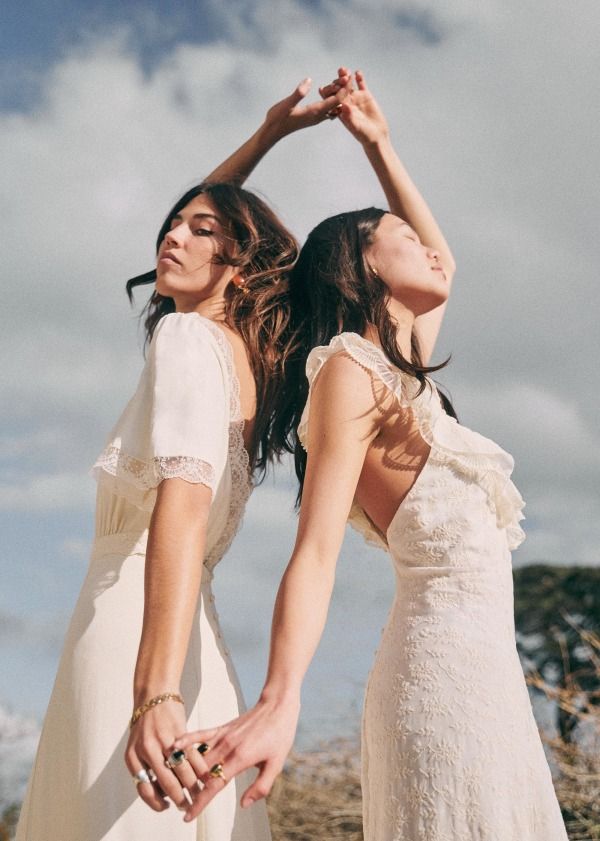 two women in white dresses are standing outdoors, with their arms around each other. they are both smiling and looking at each other, and it seems like they are happy to be together. they are both wearing simple white dresses, which makes them look like a match made in heaven. the background of the image is a beautiful blue sky with white clouds, which adds to the overall sense of happiness and peace.