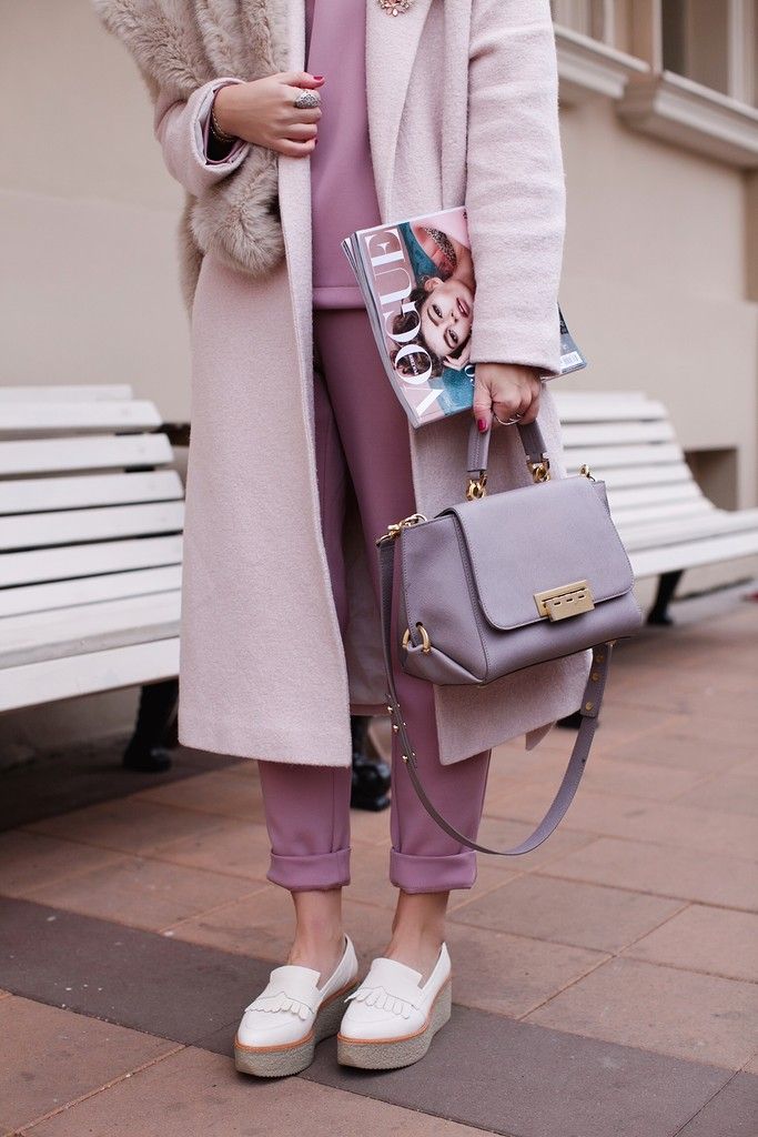the image shows a stylish woman wearing a pale pink long coat with a fur collar, mauvecolored pants, and white wedge loafers. she carries a light gray satchel and is holding a vogue magazine. the photo is taken outdoors, with a white park bench visible in the background. the overall aesthetic is sophisticated and elegant, showcasing autumn/winter fashion. the color palette is muted and pastel, emphasizing soft pink and purple tones.