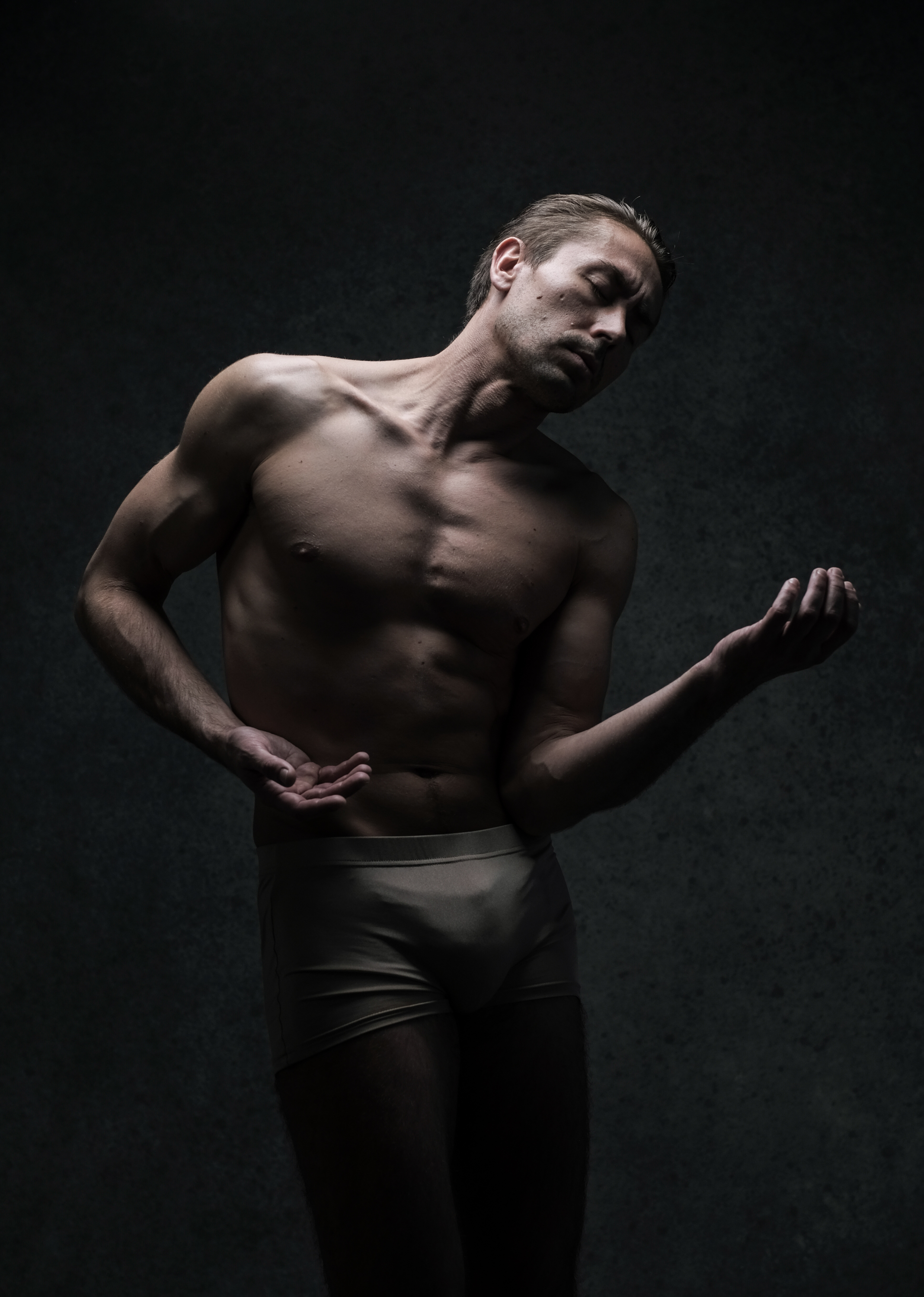 the image presents a shirtless man in underwear, striking a dance pose against a dark studio backdrop.  his physique is lean and muscular. dramatic lighting casts shadows that accentuate the contours of his body and create a sense of depth and movement. the man's head is tilted back, and his eyes are closed, conveying a sense of absorption and grace in his movement.