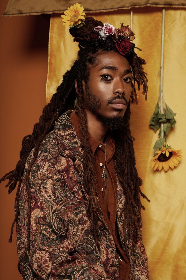a portrait of a man with dreadlocks and flowers in his hair, wearing a paisley jacket and a brown shirt with a cross necklace. he has a nose ring and eyeliner, and is looking at the camera. the background is yellow and there are sunflowers visible.