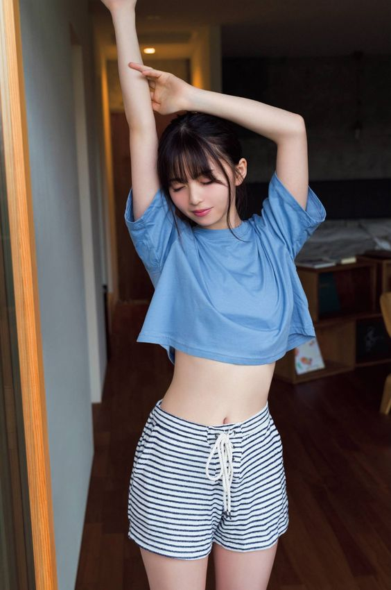 a young woman with shoulderlength dark hair, styled with bangs, stands in a doorway. she is wearing a blue cropped tshirt and black and white striped drawstring shorts. her eyes are closed as she stretches her arms above her head, her right hand resting on her left wrist. the room behind her is minimally furnished with a wooden floor and a glimpse of a bed and shelves. the lighting is bright and soft, illuminating the scene with a natural feel.
