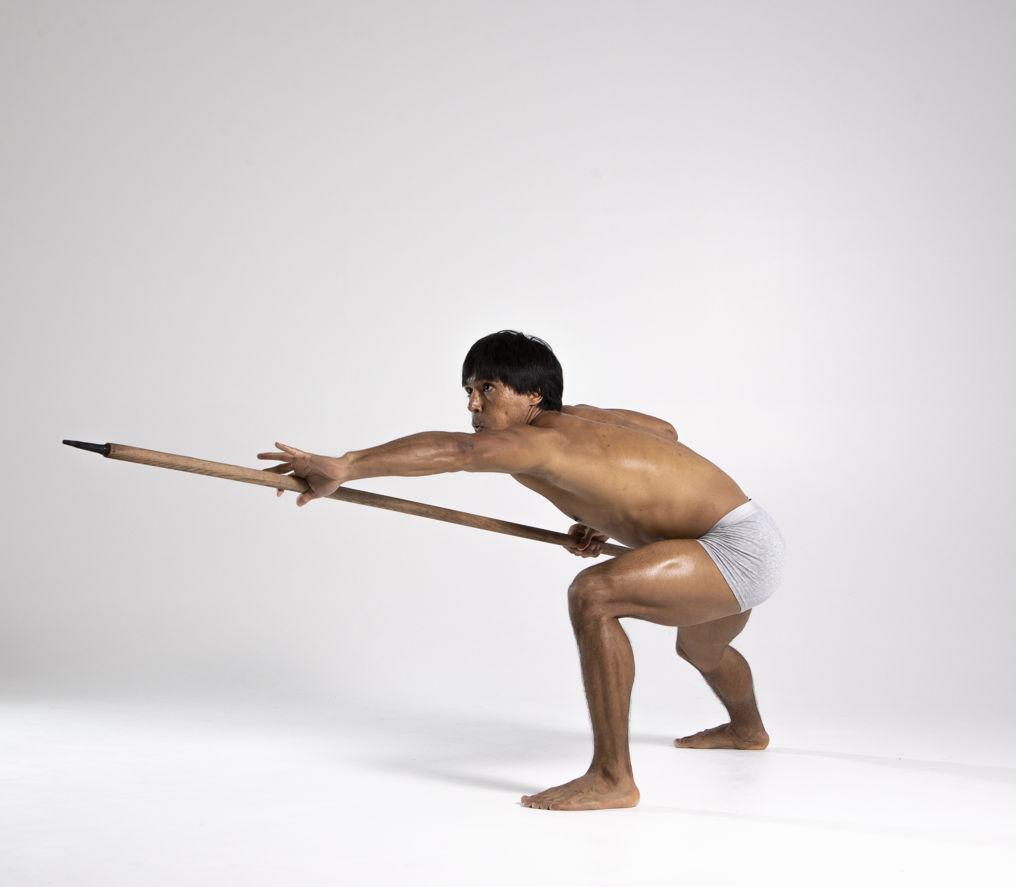 a barechested man wearing a loincloth crouches in a fighting stance, aiming a spear forward. he has dark hair and an athletic build, with visible muscles. his posture is defensive and alert, suggesting a primitive warrior preparing for an attack. the background is plain white, emphasizing the figure and weapon.