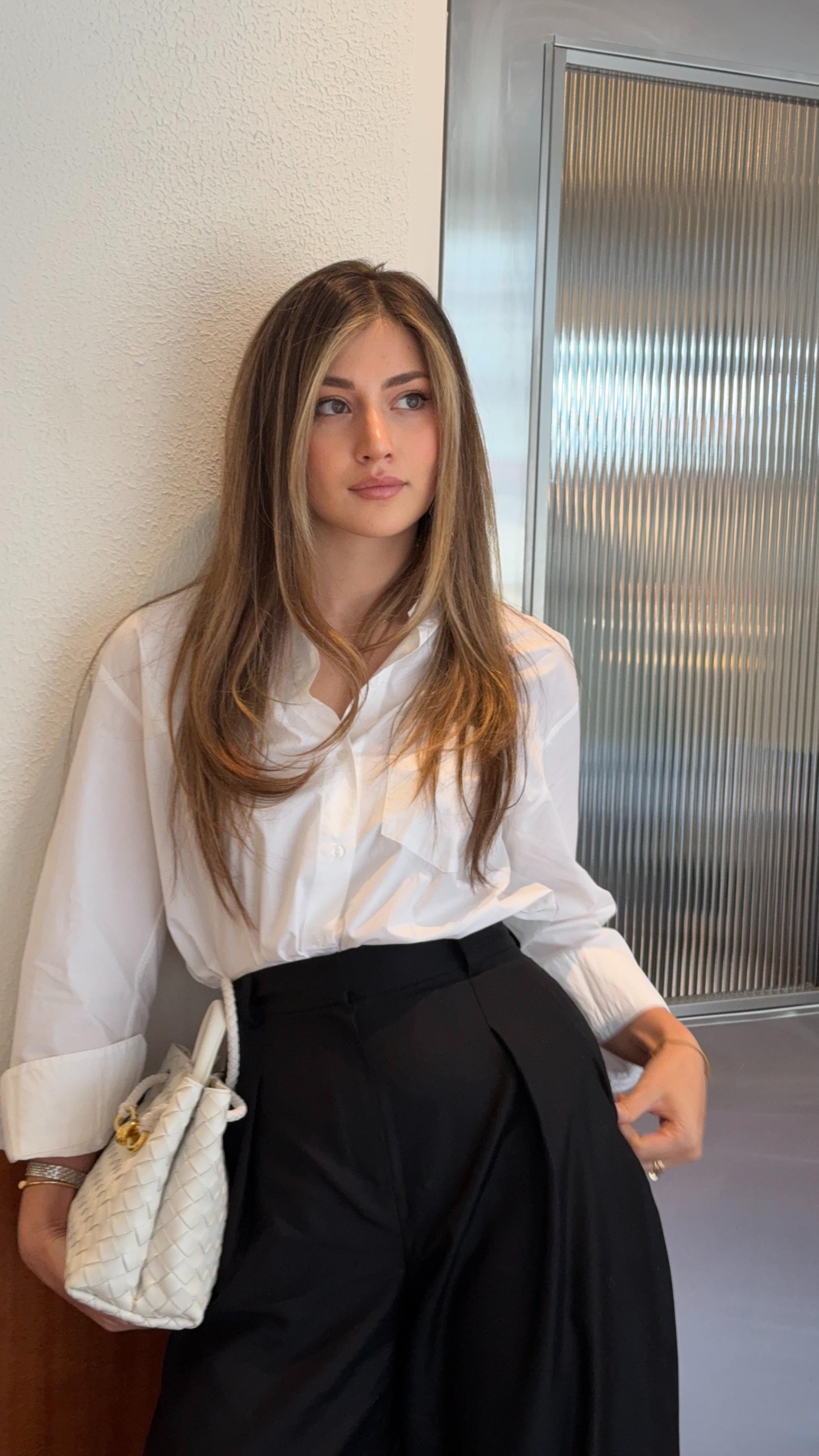 a stylish woman is posing for a photo. she is wearing a white buttondown shirt and black pants. she is carrying a white handbag. the woman has long, blonde hair. she is wearing jewelry on her wrist and fingers. the background features a white wall and a textured glass door.