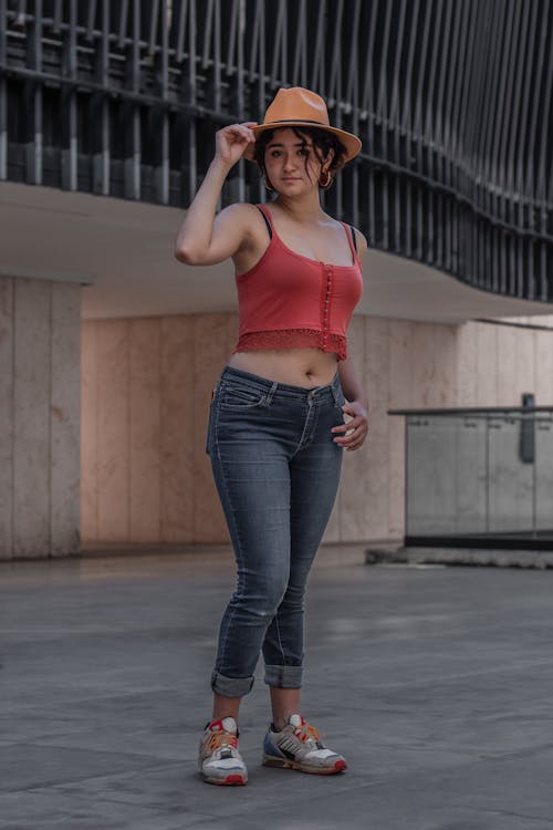 a young woman stands confidently outdoors in a stylish outfit. she's wearing a coral crop top, dark wash jeans cuffed at the ankles, and a light brown fedora hat.  her shoes are light gray and multicolored sneakers. the background features a modern building with a unique metal facade.  the overall impression is one of relaxed urban style.