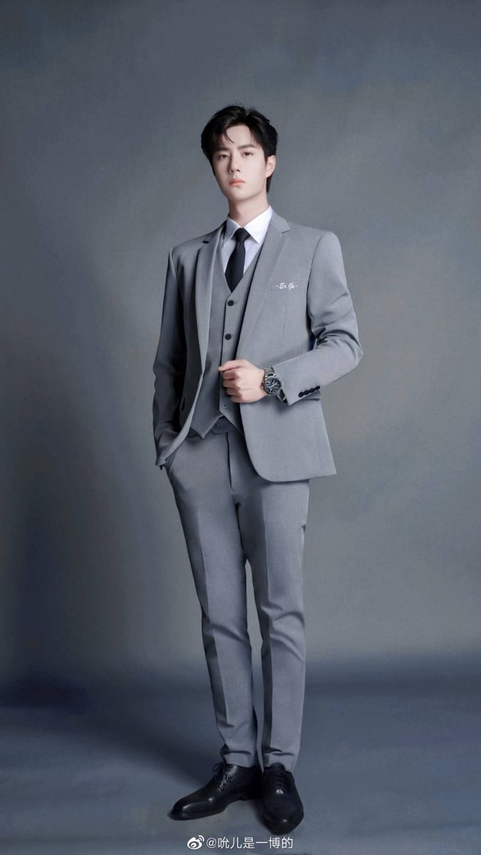 a fullbody studio shot of an asian man wearing a grey suit with a black tie and vest. he is standing with one hand in his pocket and wearing a wristwatch. his dress shoes are black. he is wearing formal wear.