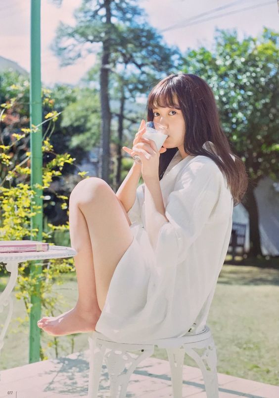 a young woman with dark hair and bangs sits on a white metal chair on a patio. she is wearing a loose white dress and is barefoot. she is drinking milk from a clear glass and is looking directly at the viewer. to her left is a small white table with a book resting on top. the scene is bathed in bright sunlight and features lush greenery in the background, evoking a sense of summery relaxation.