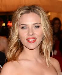 a closeup portrait of scarlett johansson. she has shoulderlength, wavy blonde hair and is wearing red lipstick.  the background is blurred but appears to be a red carpet event.  she is looking directly at the camera.