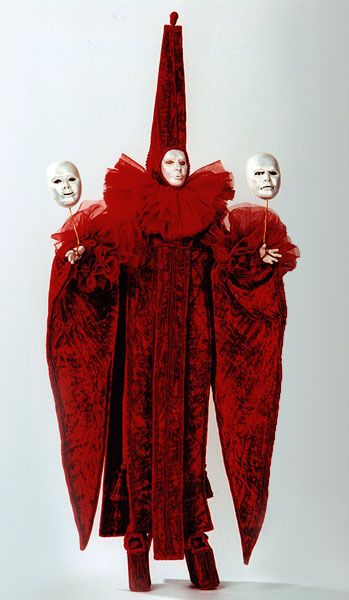 a person in a red costume is holding two white masks in front of them. they are in a standing position with one mask held on their left and the other on their right. the person is wearing a long robe like costume that has a large ruffle around the neck. the costume has a very dramatic and theatrical feel.