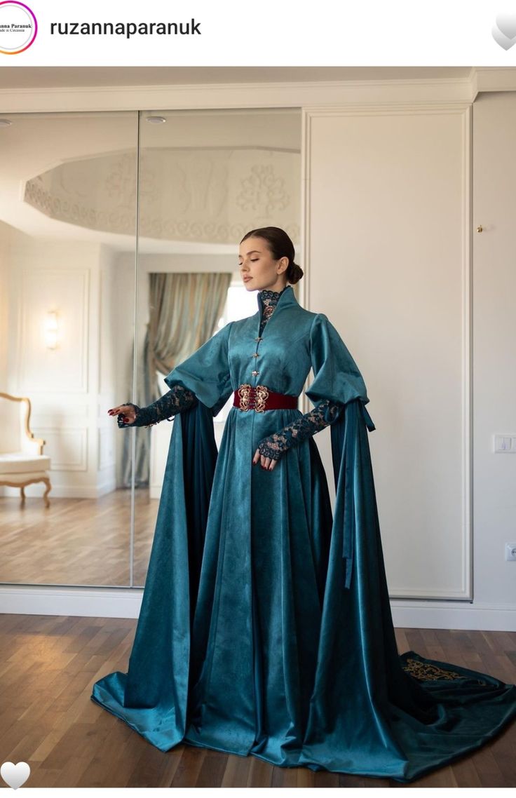 the image showcases a model wearing a stunning teal velvet gown. the dress features long, flowing sleeves, a high neck, and an ornate, dark red belt accentuating the waist. the fabric drapes elegantly, creating a flowing silhouette with a long train that pools on the floor. the model stands in a grand, ornate interior setting with mirrors reflecting the scene.  the overall style is regal and elegant, hinting at a vintage or historical aesthetic. the image appears to be professional fashion photography.