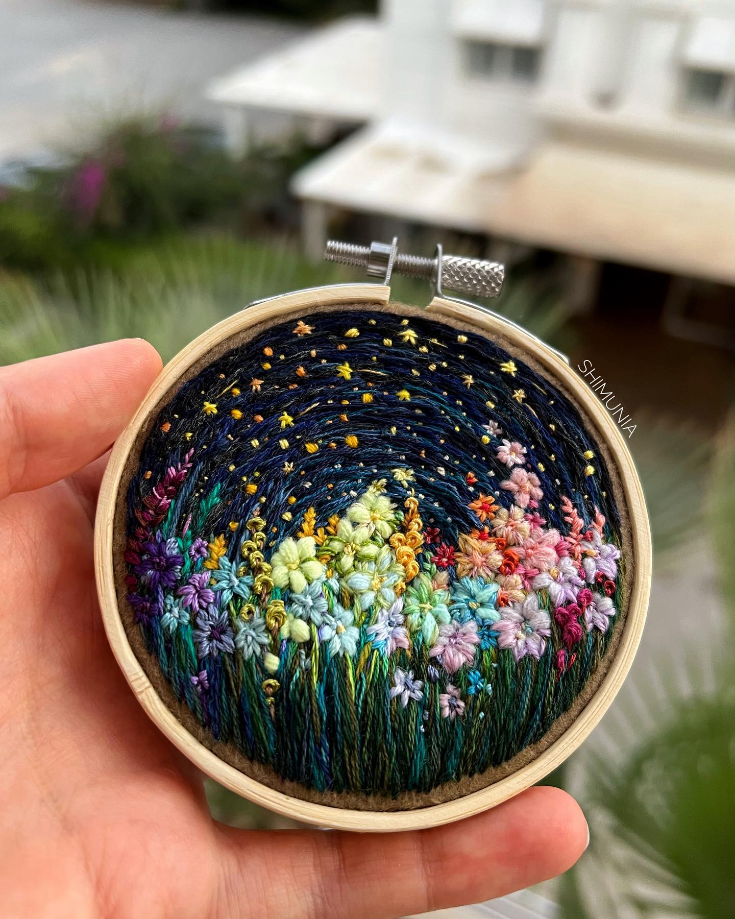 a close up image of a person holding an embroidery hoop with their left hand. the embroidery hoop contains a work of art featuring a field of colorful flowers under a navy blue night sky filled with yellow stars.
