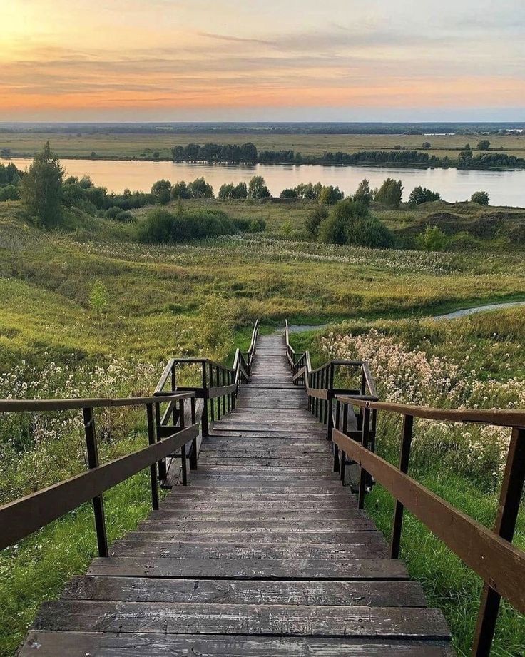 the image shows a long wooden staircase descending a grassy hill towards a river. the staircase has railings on both sides. the surrounding landscape consists of a green field with wildflowers, a river with trees along its banks, and a sunset sky. the view is from above, looking down the staircase.