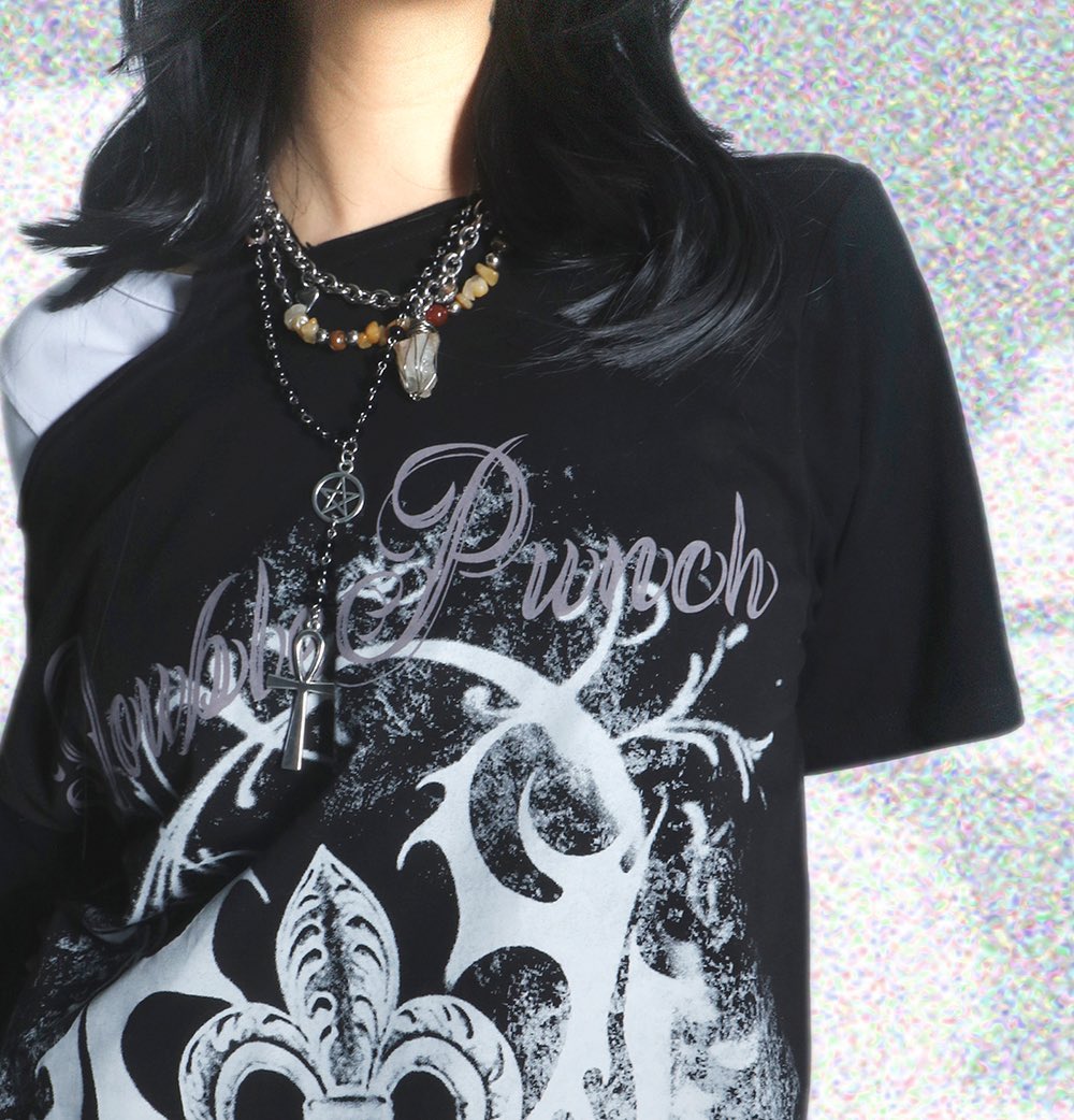 the image shows a closeup of a person wearing a black tshirt featuring a distressed graphic design with the words "purple punch" incorporated into the design. the design has gothic/metal aesthetics.  the person is wearing multiple layered necklaces with various pendants, including what appears to be an anubis pendant and a pentagram pendant, along with other stones and crystals.  the person has long, dark hair, and the overall style is dark and alternative. the background is blurry and iridescent.