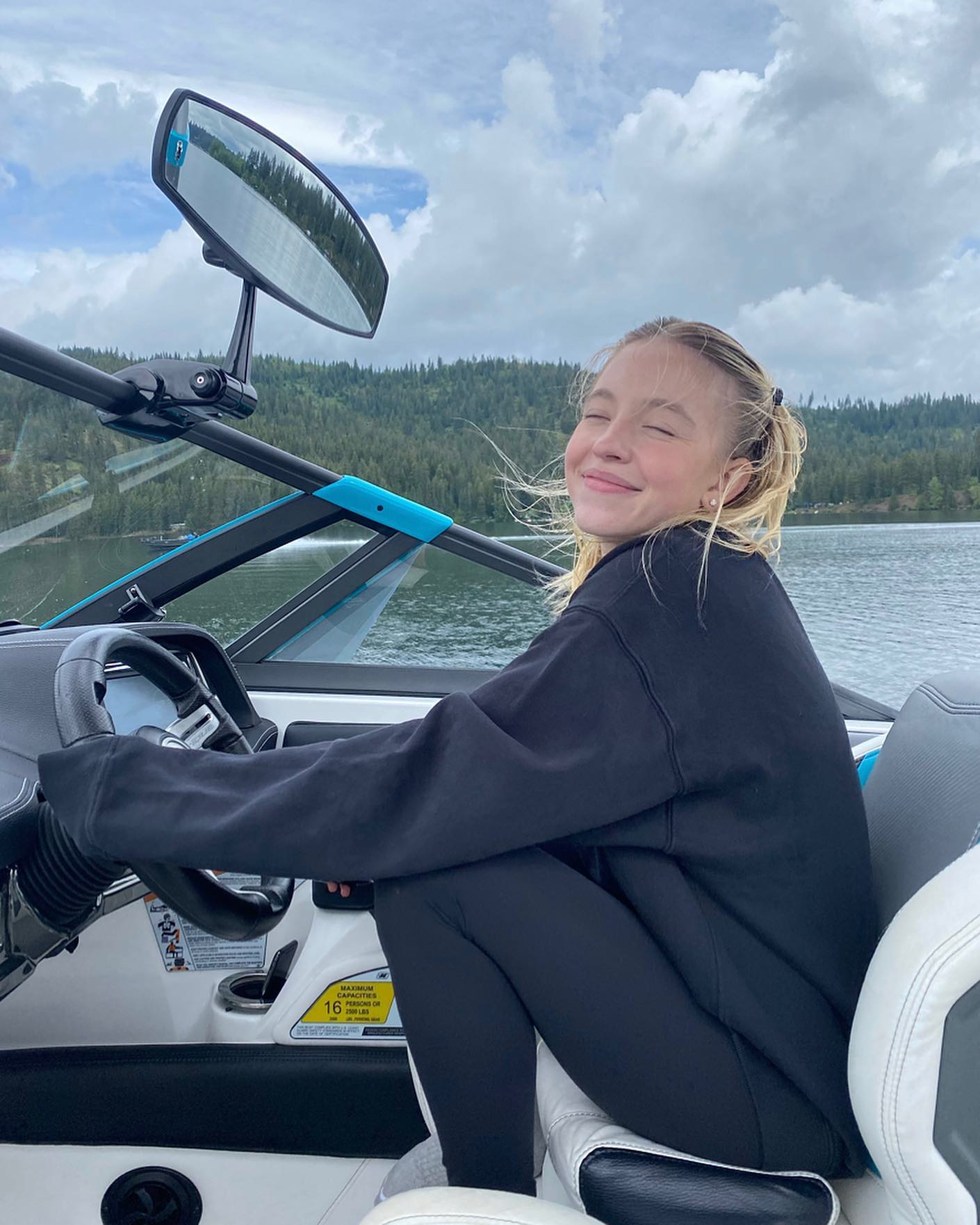 a young blonde girl sits happily in the driver's seat of a boat on a sunny summer day.  she's smiling and appears to be enjoying herself. the boat is on a lake surrounded by lush green mountains and trees under a partly cloudy sky. the girl is wearing a black sweatshirt and black leggings. the overall scene is one of summer leisure and carefree fun.