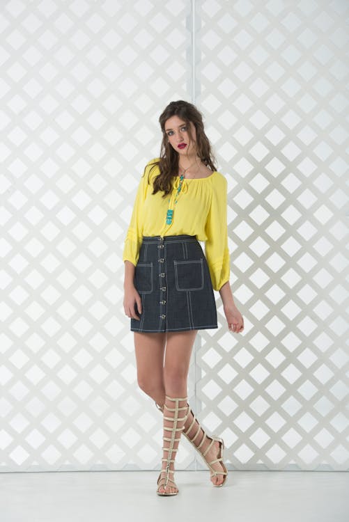 a woman stands against a white lattice background, dressed in a vibrant yellow, longsleeved blouse paired with a dark denim mini skirt featuring visible buttons and front pockets. she accessorizes with a blue turquoise necklace. her footwear consists of beige gladiator sandals that extend up to her knees.