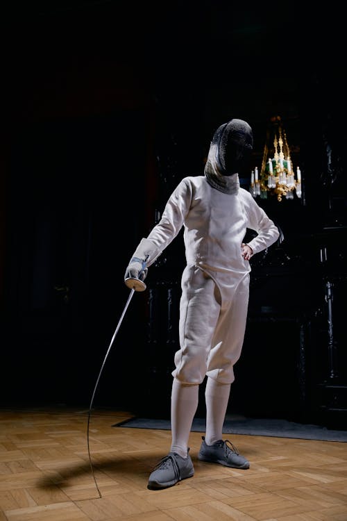 a fullbody shot of a fencer in a white uniform and mask stands on a wooden floor. the fencer holds a foil and strikes a poised stance in a dark room with a partially visible antique piece of furniture and chandelier in the background. the image is sharply focused, showing details of the uniform and equipment.  the lighting enhances the dramatic effect.