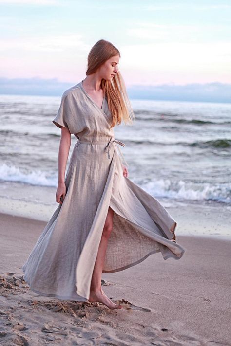 a young woman with long blonde hair is standing on a sandy beach wearing a flowing, light gray linen wrap dress. the dress has a vneckline, short sleeves, and a slit on one side. the skirt of the dress is billowing in the gentle breeze. the ocean is in the background, and the sky is a soft pink and blue. the photo was taken at eye level in soft, natural light.