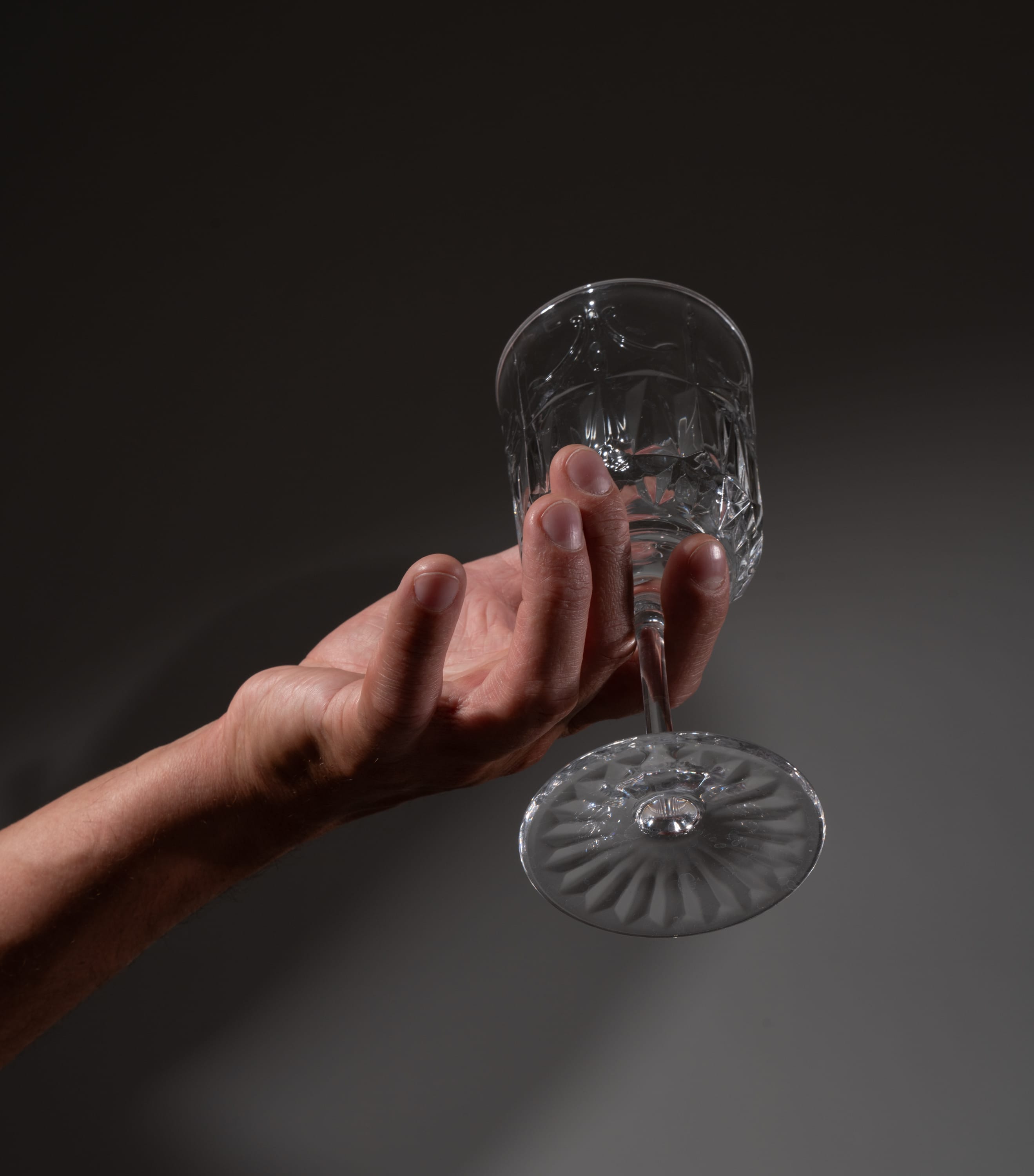 a hand delicately holds an empty crystal wine glass against a muted gray backdrop. the perspective is from below, emphasizing the fragility and elegance of the glass.