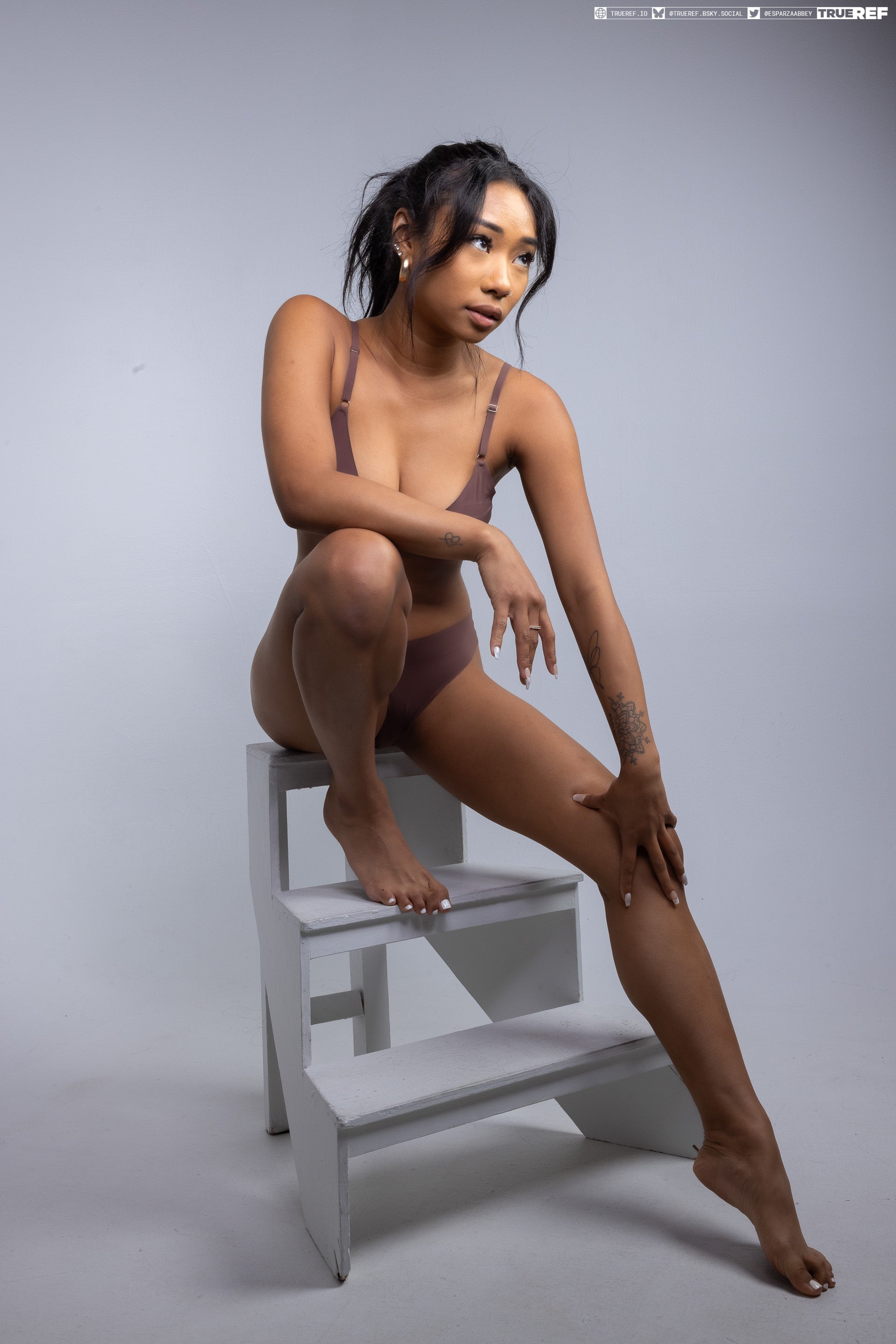 a woman in a bra and underwear is sitting on a stool in a studio. she has a tattoo on her arm and is barefoot.