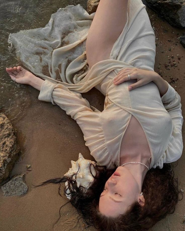 a woman is lying on a sandy beach next to the ocean, wearing a lightcolored dress. her curly hair is spread around her head, and a seashell is placed near her head. she is wearing a necklace and a ring. the scene is bathed in soft, natural light, with rocks visible on the left side of the frame. the edge of the water is visible near the top of the image.