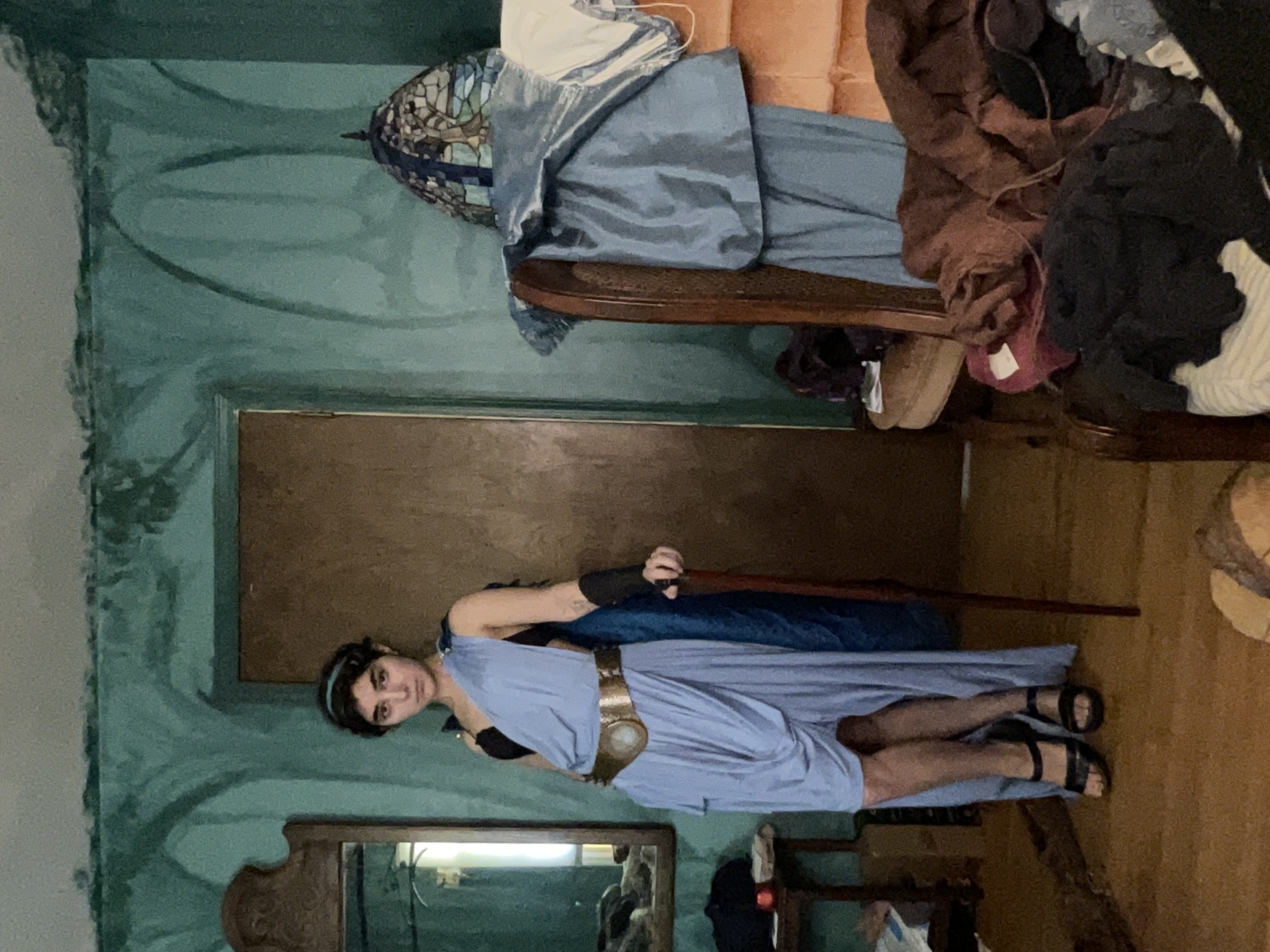 a fullbody shot of a young woman posing in what appears to be a homemade ancient greek or roman goddess costume. she's wearing a light blue, highlow dress with a gold belt and dark sandals.  her accessories include a headband and dark arm coverings. she holds a wooden cane.  the background is a vintagelooking room with teal walls, an antique mirror, and a wooden chair piled with clothes. the overall impression is that the photograph is a cosplay portrait taken in a personal space.