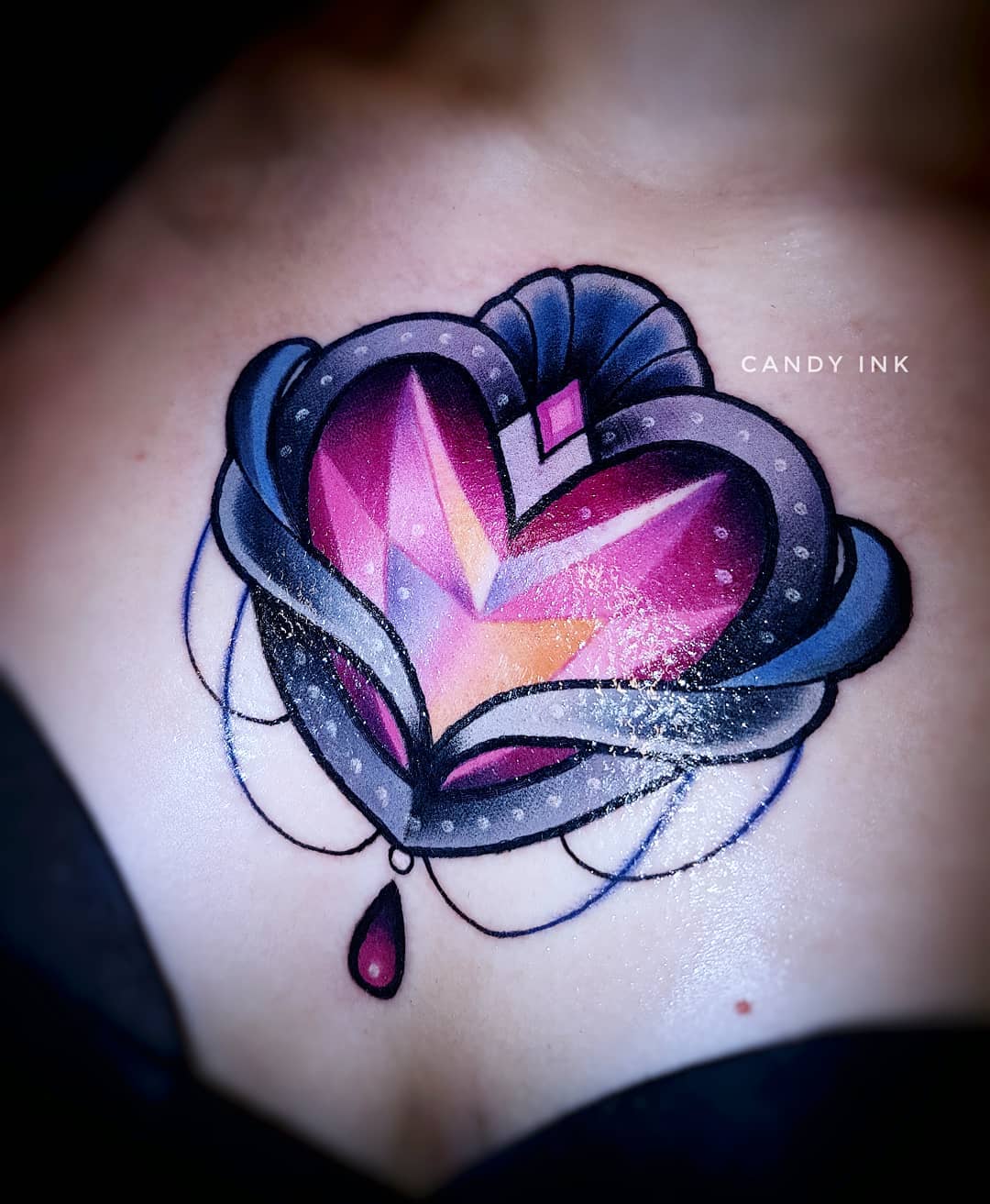 a closeup photograph showcasing a colorful neotraditional tattoo of an ornate heartshaped gemstone on someone's chest. the heart is rendered in shades of pink and purple, encased in a stylized silver locket adorned with swirling details and a single dangling teardrop. the tattoo is executed in a bold and graphic style with clean black outlines.