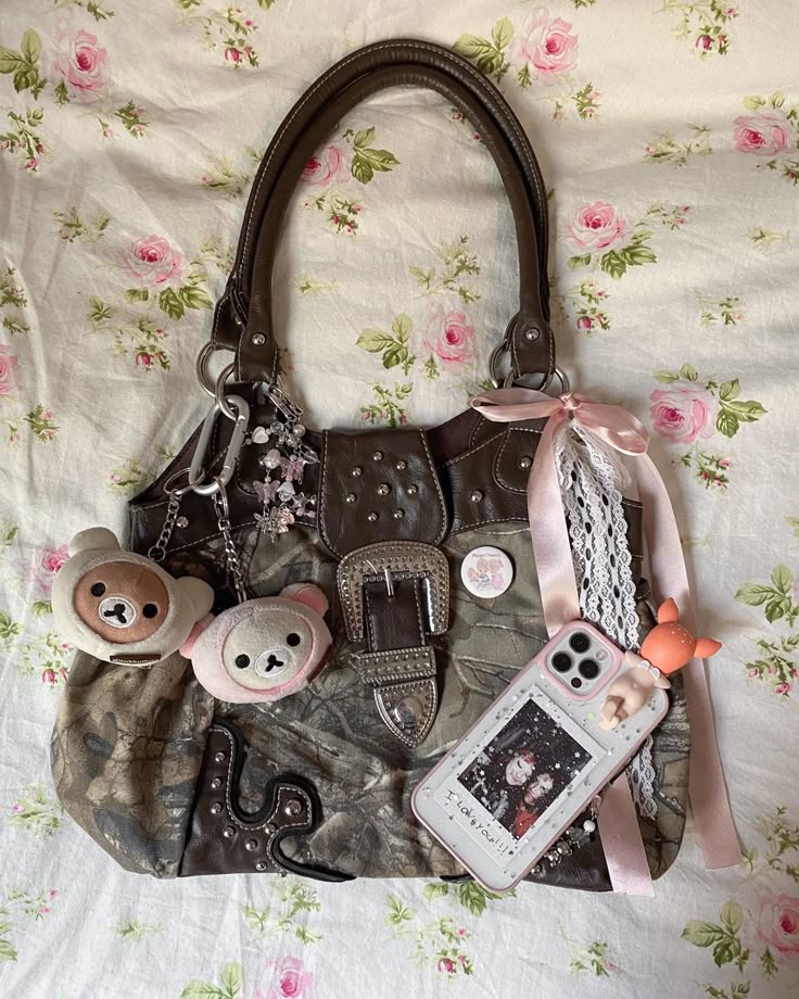 a closeup overhead shot of a decorated handbag on a floral background. the handbag is adorned with various charms, including plush bear keychains, a phone case, ribbons, lace, and a silver buckle. the background is a floral pattern with pink roses and green leaves on a creamcolored fabric.
