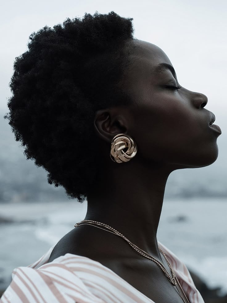 a profile portrait of a black woman with closed eyes, looking upward. she has short, dark, afrotextured hair, and is wearing a large gold knotstyle earring and a layered gold necklace. the background is blurred, suggesting a beach environment with water and possibly rocks. she is wearing a striped top. the lighting is soft and natural, emphasizing the smoothness of her skin. the overall mood is one of peace and contemplation.