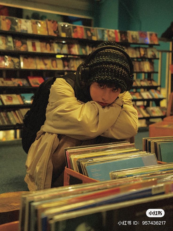 a young asian woman sits in a record store, surrounded by shelves of vinyl records and cds. she's wearing a knitted beanie, headphones, and a beige jacket. she has a pensive expression and is looking directly at the camera. the image is dimly lit, giving it a moody atmosphere. the focus is on the woman, with the records in the background slightly blurred.