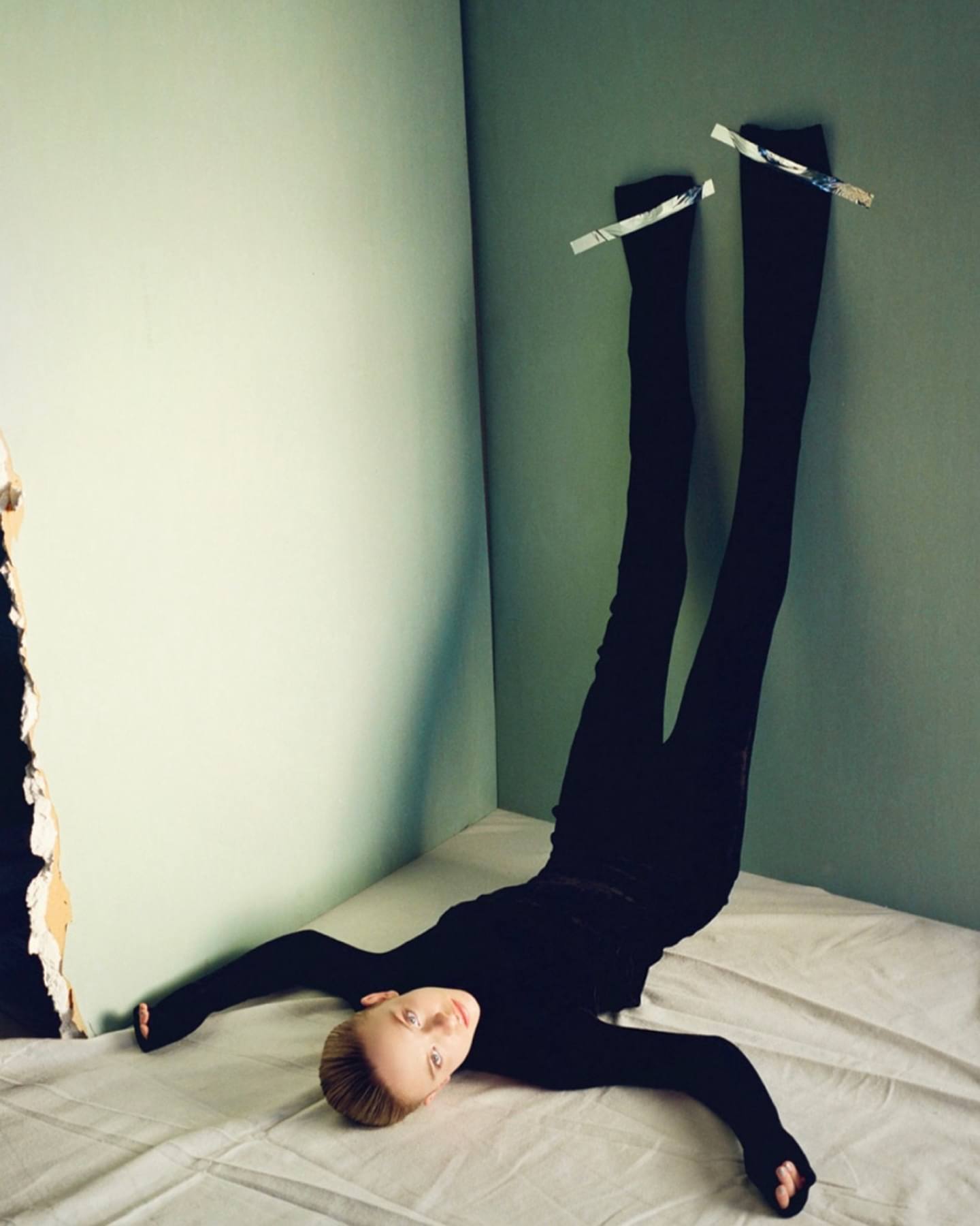 fashion photograph of a slender woman wearing all black clothing. her legs are extended upward along a wall. the model is laying on the floor, which appears to be a studio setting. the style is minimalist.