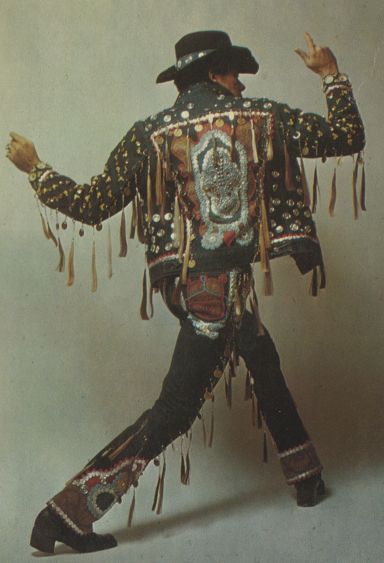 a man is wearing a black jacket with fringes and dark blue jeans.  the jacket is decorated with silver studs and beads. the man has a black hat on and is posing with his right hand raised in the air. the image is in a studio setting. the photo is likely from the 1960s or 1970s. he is a musician, possibly a rock and roll singer or guitarist.