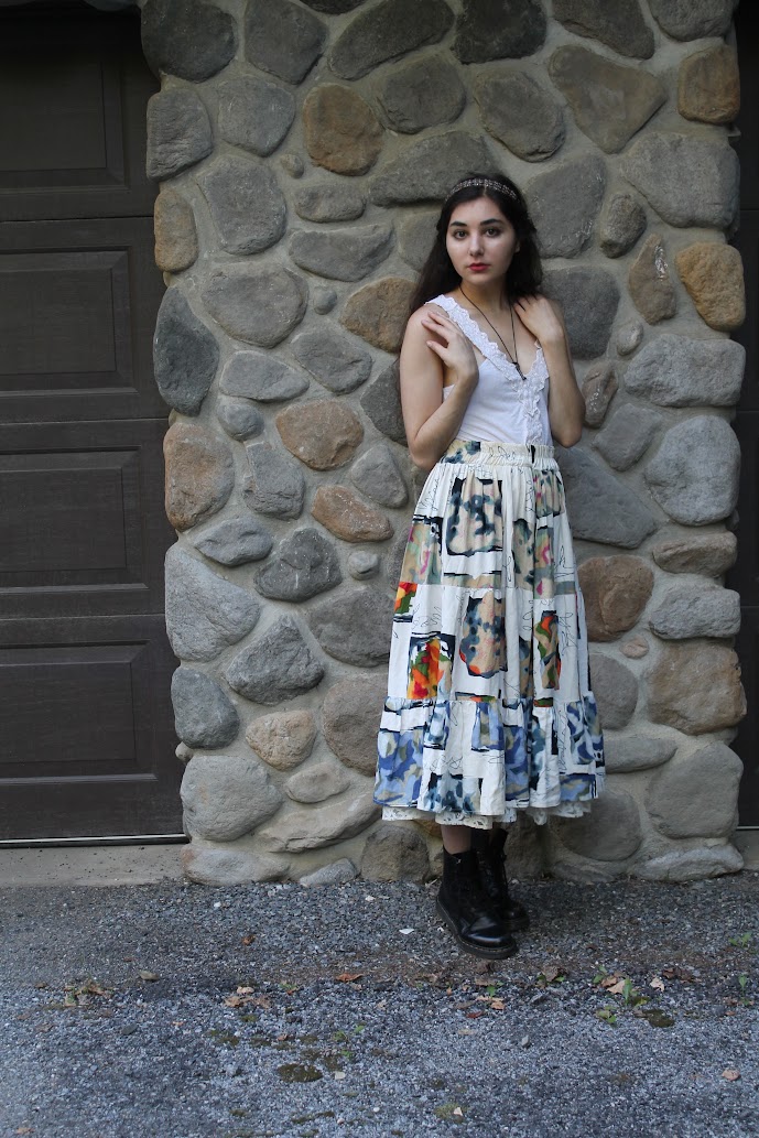 a young woman stands against a stone wall wearing a long, patchwork skirt and a white lacetrimmed tank top. she has dark hair, a headband, and is wearing black boots. she is looking at the camera. the style is bohemian or hippie.