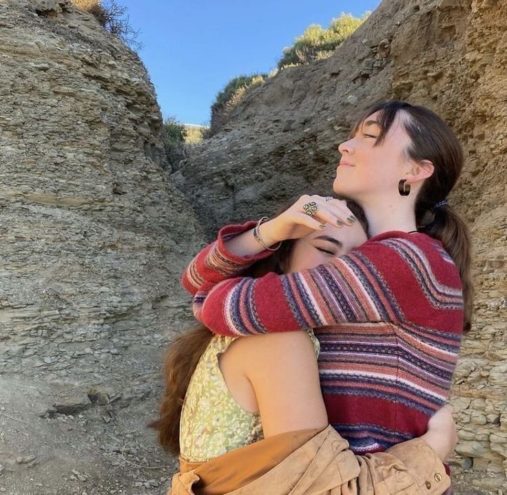 two women are hugging each other with their eyes closed while standing in front of a rocky cliff. the woman on the right is wearing a red and white striped sweater, while the woman on the left is wearing a yellow floral dress. they are both smiling, and their faces are illuminated by the sun. they look happy and in love. the rocky cliff is in the background and a lot of green bushes can also be seen.  this image is warm, happy, and romantic. it shows the love and care between two people.