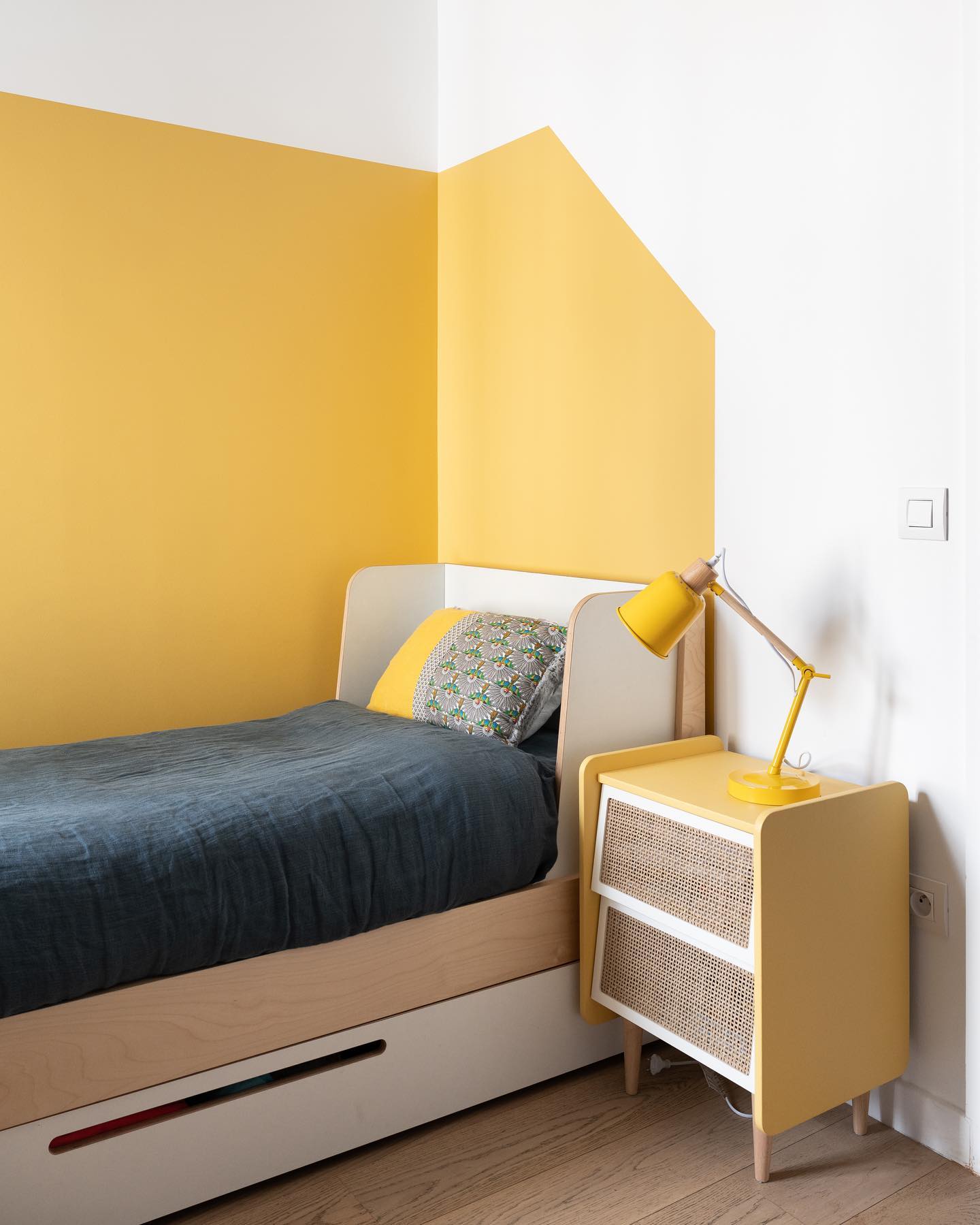 a bright and modern child's bedroom features a single bed with a white frame and light wood accents. the bed is covered with a dark blue duvet and has a patterned pillow. a yellow and white wall is behind the bed. a yellow nightstand with woven drawers stands next to the bed, topped with a yellow desk lamp. the room has hardwood floors and a minimalist design. a light switch and electrical outlet are visible on the white wall.