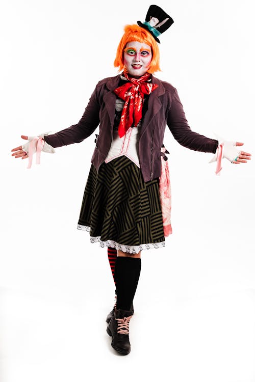 a fullbody shot of a woman dressed in a mad hatter costume from alice in wonderland. she has bright orange hair, a top hat, a dark jacket, and a patterned skirt. her makeup is styled to resemble the mad hatter's character, with exaggerated features. she is posed with her arms outstretched, and her expression is playful and whimsical. the background is plain white, making the costume and the model the focus of the image. the photograph appears to be a professional studio shot, possibly for a theatrical production or cosplay event.