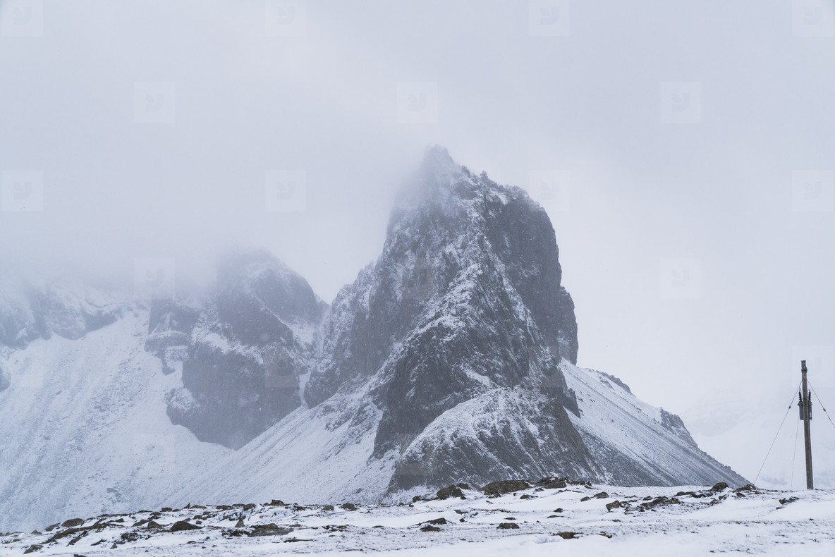 a tall, rocky mountain peak rises up from a snow covered landscape. the mountain is shrouded in fog and clouds. the sky is a pale gray and the ground is covered in snow. the landscape is desolate and cold.