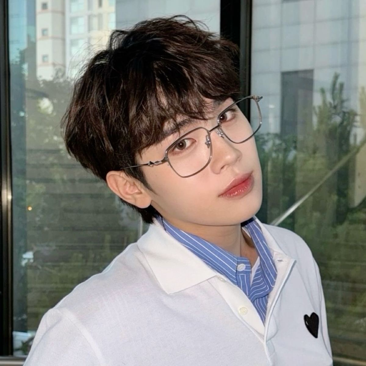 the image shows an asian man with short, dark hair wearing glasses. he is wearing a white collared shirt with thin blue stripes. he is posing for a selfie indoors, with a window behind him. the background is blurred, and the lighting is natural.